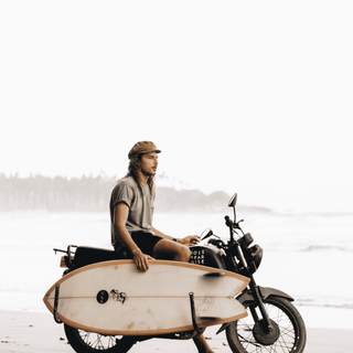 Man with surfboard sitting on a motorcycle wearing Will & Bear Baker Tan cap.