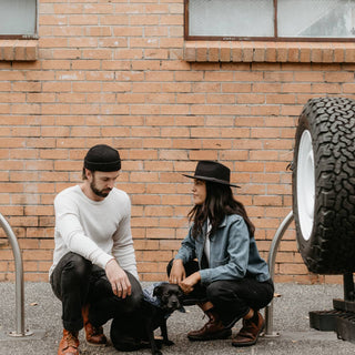 Urban scene featuring woman wearing Will & Bear Calloway Black Wide Brim Hat, sitting with man and dog against brick wall