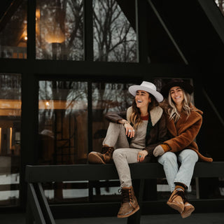 Two women outside a cabin wearing Will & Bear fedoras, featuring the Cooper Bone White Upturned Fedora in a winter setting.