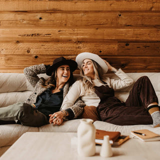Couple lounging indoors in winter wearing Will & Bear fedoras, including the Cooper Bone White Upturned Fedora.