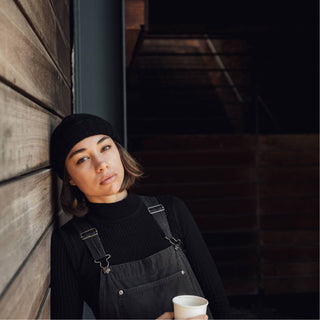 Woman leaning against wooden wall, wearing the Will & Bear Doc Black Wool Fisherman’s Beanie and holding a coffee cup.