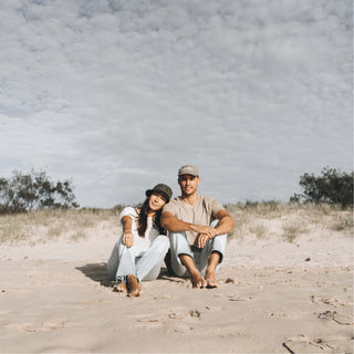 Couple sitting on the sand at the beach, both wearing Will & Bear Earth Bucket Organic Hemp Bucket Hats.