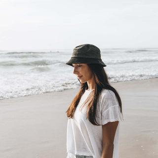 Woman standing on the beach in the Will & Bear Earth Bucket Organic Hemp Bucket Hat, with ocean waves in the background.