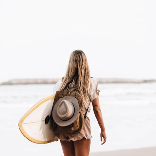 Woman walking on beach with a surfboard and wide brim hat clipped to her backpack using Will & Bear Hat Carry Clips.
