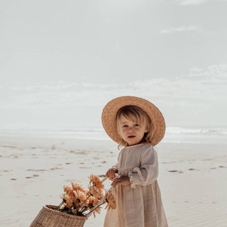 Toddler Wearing Will & Bear Little Harvey Amber Kids Straw Hat – Holding Basket of Flowers on the Beach.